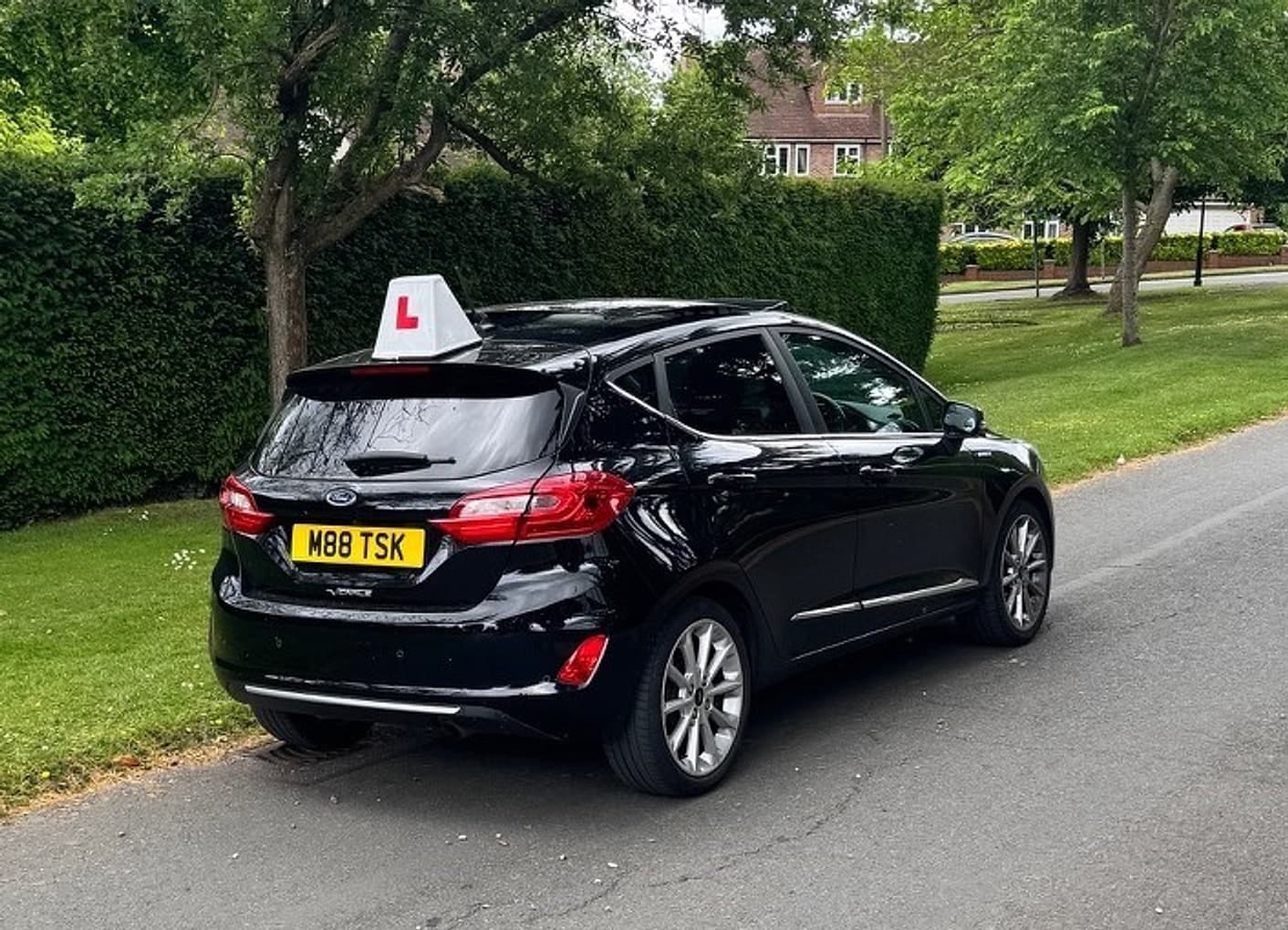 TF Driving’s black automatic tuition car with an L-plate roof sign, parked beside a green on a residential road