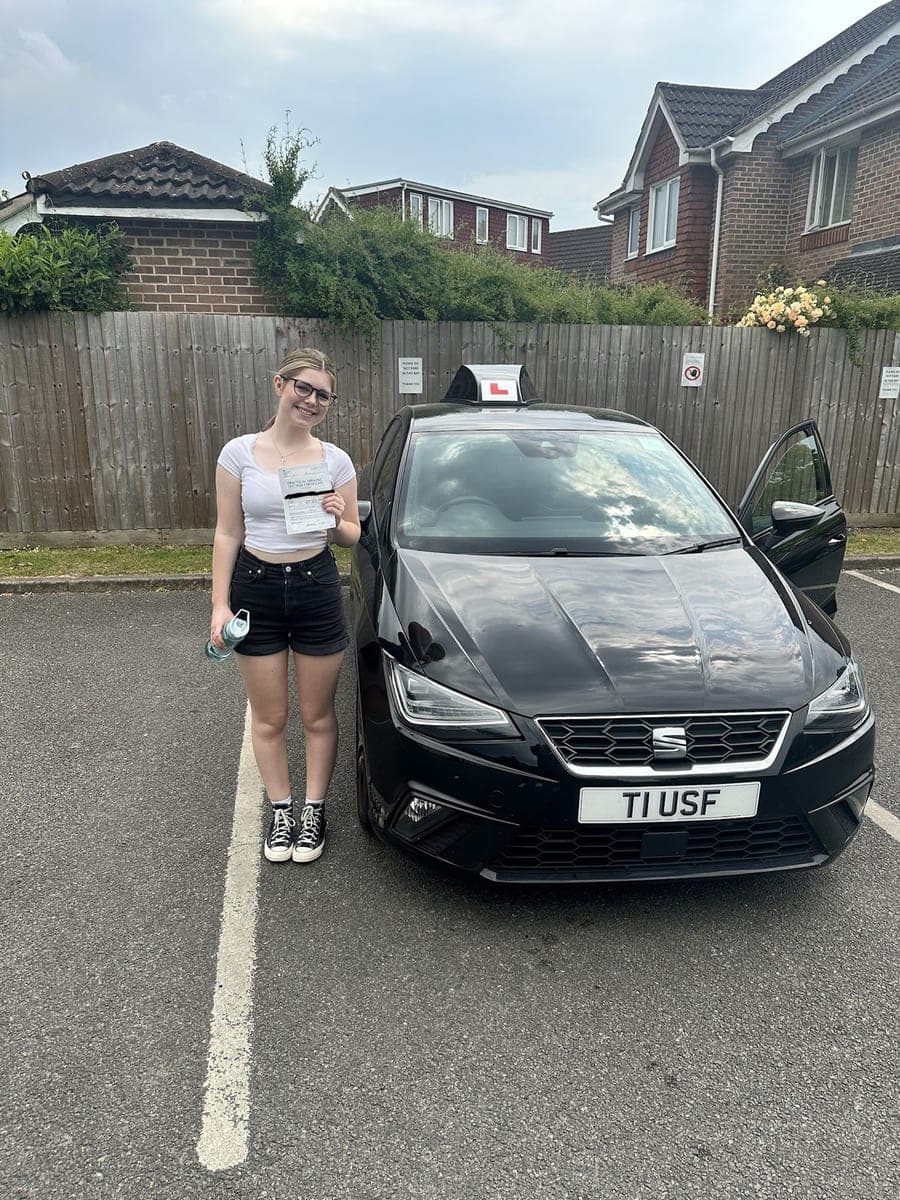 A smiling TF Driving pupil in glasses holding up her pass certificate next to the tuition car