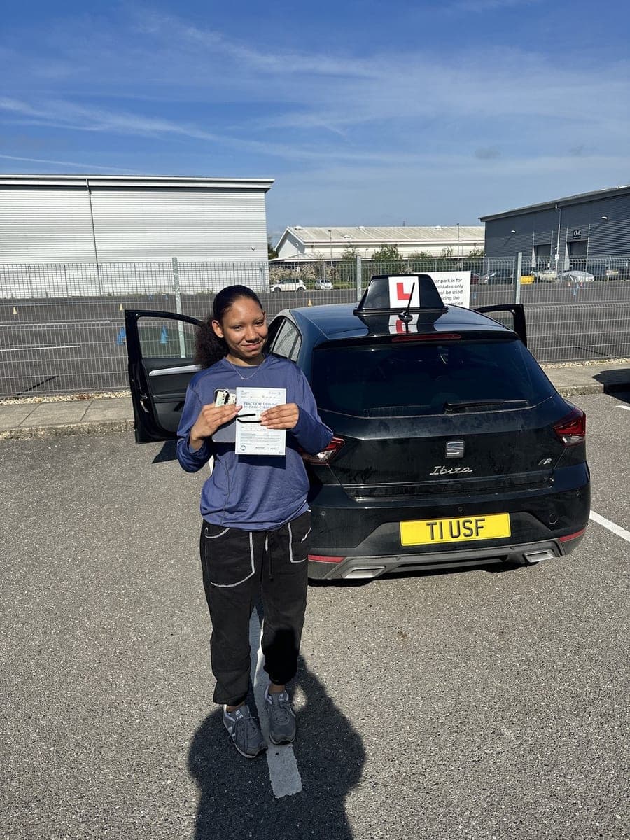 A TF Driving pupil holding her pass certificate by the open door of the tuition car on a sunny day